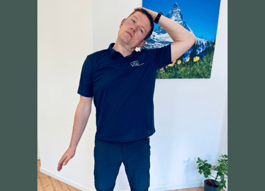 Ein Mann steht aufrecht in einem Raum und führt eine Nackendehnungsübung durch. Er trägt ein dunkelblaues Polo-Shirt mit dem Logo „Chemnitz 