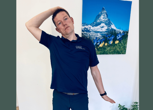 Ein Mann steht aufrecht in einem Raum und führt eine Nackendehnungsübung durch. Er trägt ein dunkelblaues Polo-Shirt mit dem Logo „Chemnitz 